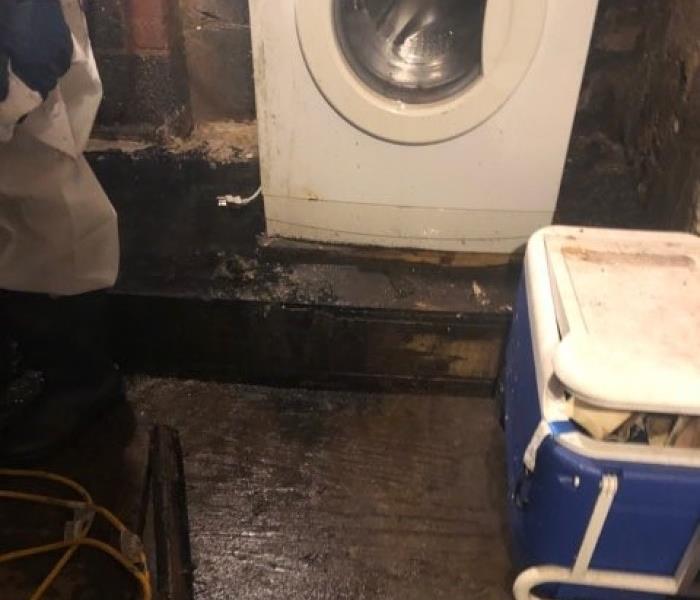 Basement wood floor, half stained black due to flood.  A pool of water is still visible next to a washing machine.
